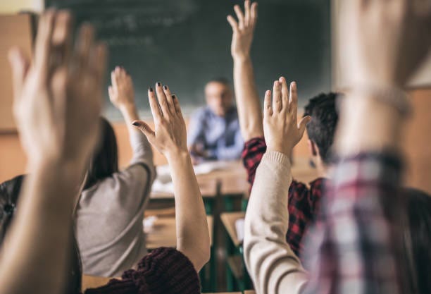 Five people sitting with the hands raised in a class-room setting. There’s a teacher sitting at the front of the class with a black-board behind him.