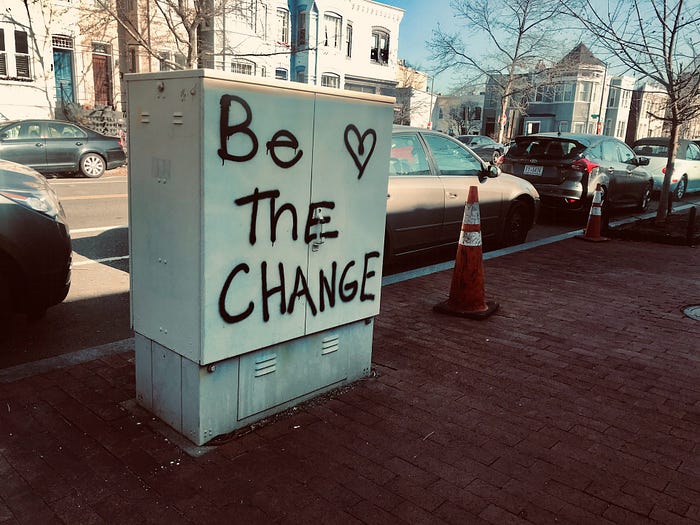 The phrase Be The Change with a heart spray-painted in black onto the side of an off-white mechanical box on a sidewalk