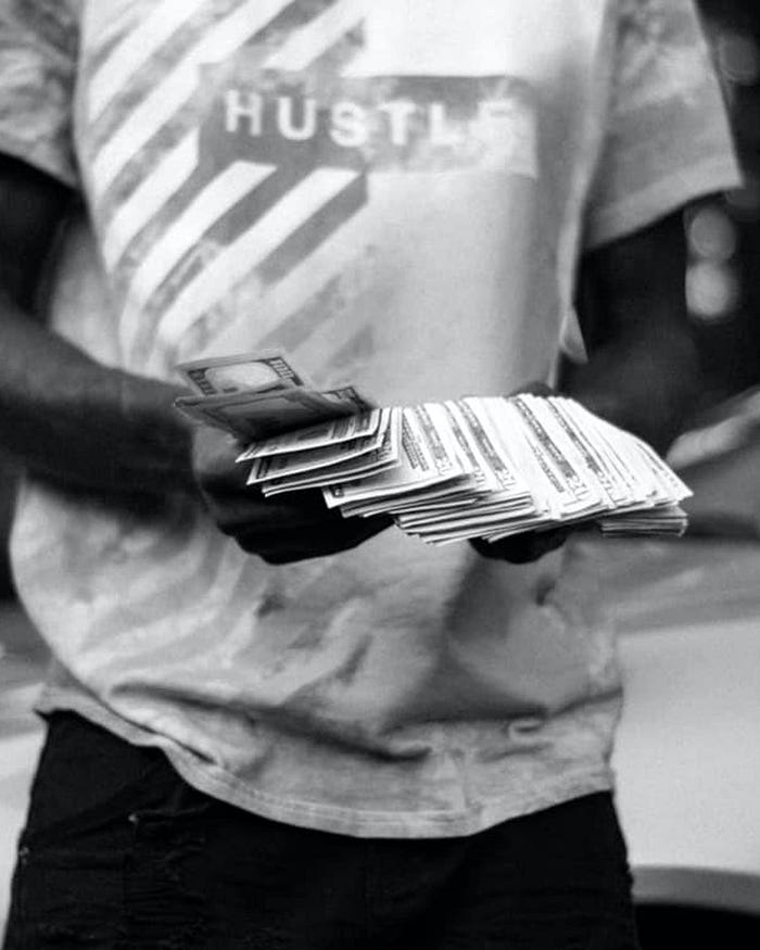 A man is wearing a white-t shirt with the word Hustle on it. He is counting a wad of money spanned out in his hands.